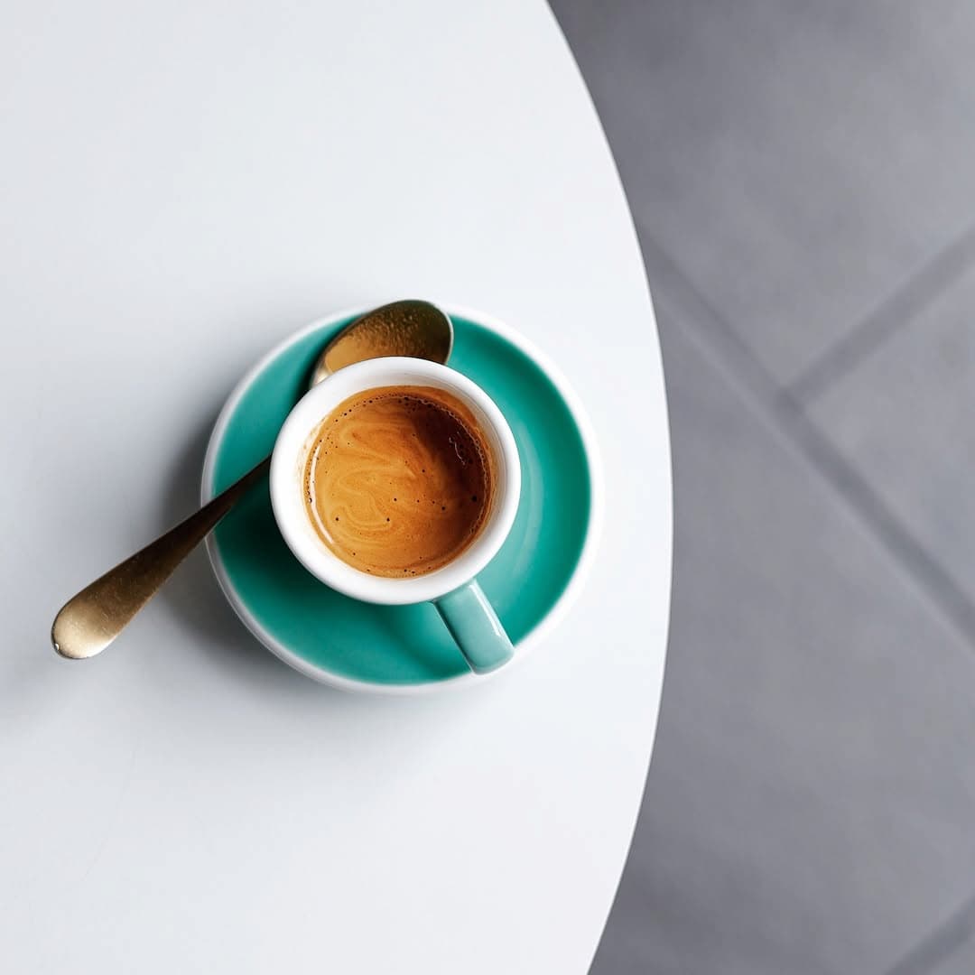 Espresso shot with a spoon on a saucer on a white surface.
