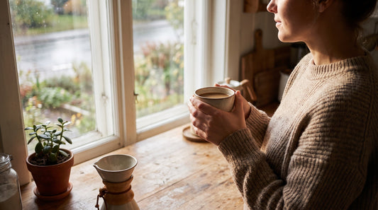 Person holding a warm mug of coffee at a kitchen counter in morning light, illustrating the protein coffee vs mushroom coffee comparison