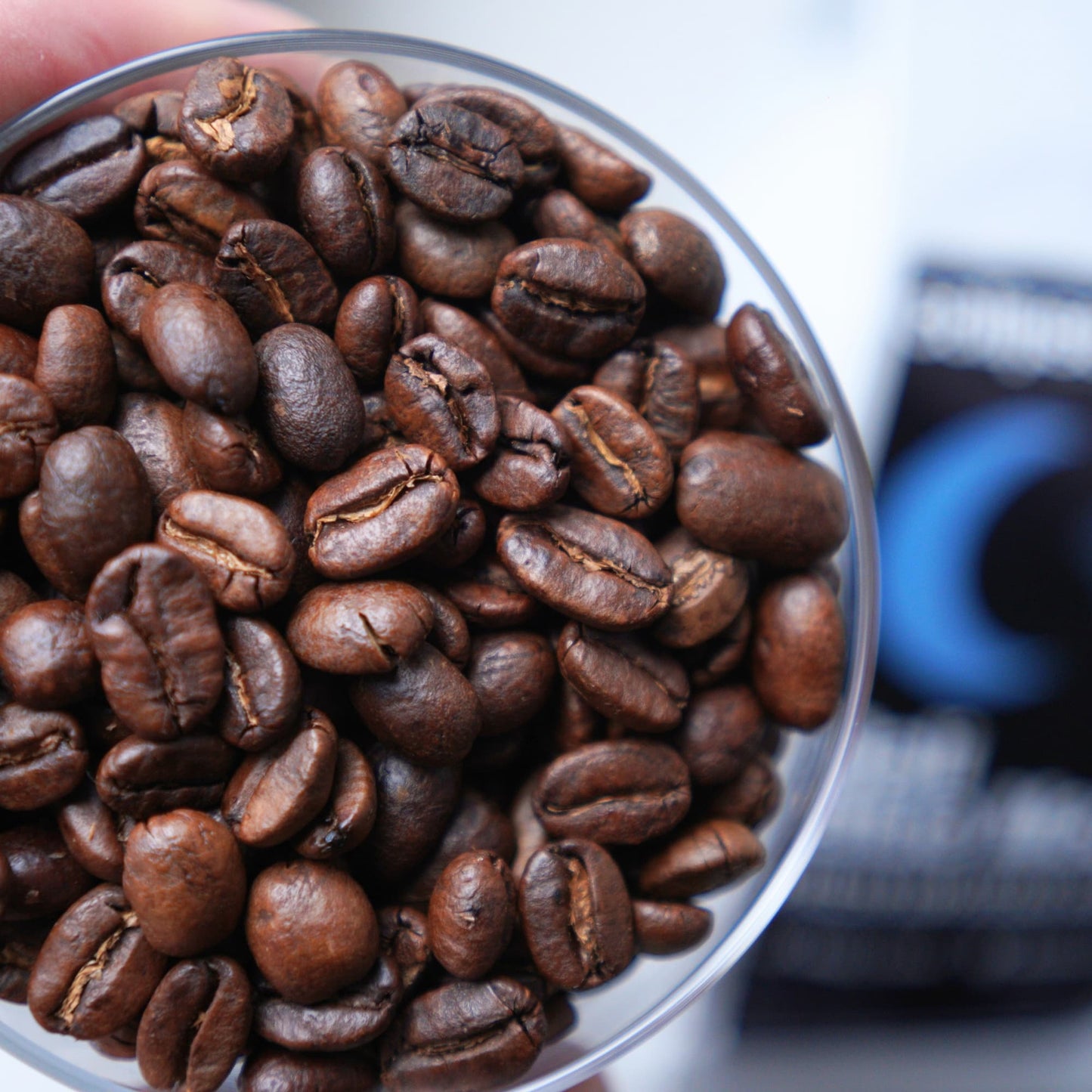 Close-up of coffee beans in a clear container with a blurred coffee package in the background.
